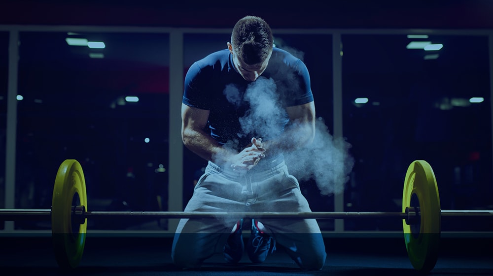 A man kneeling and rubbing powder into his hands before lifting a barbell at a gym